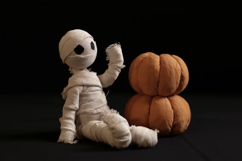 Handmade mummy figurine wrapped in white gauze bandages beside a plush orange pumpkin on black background