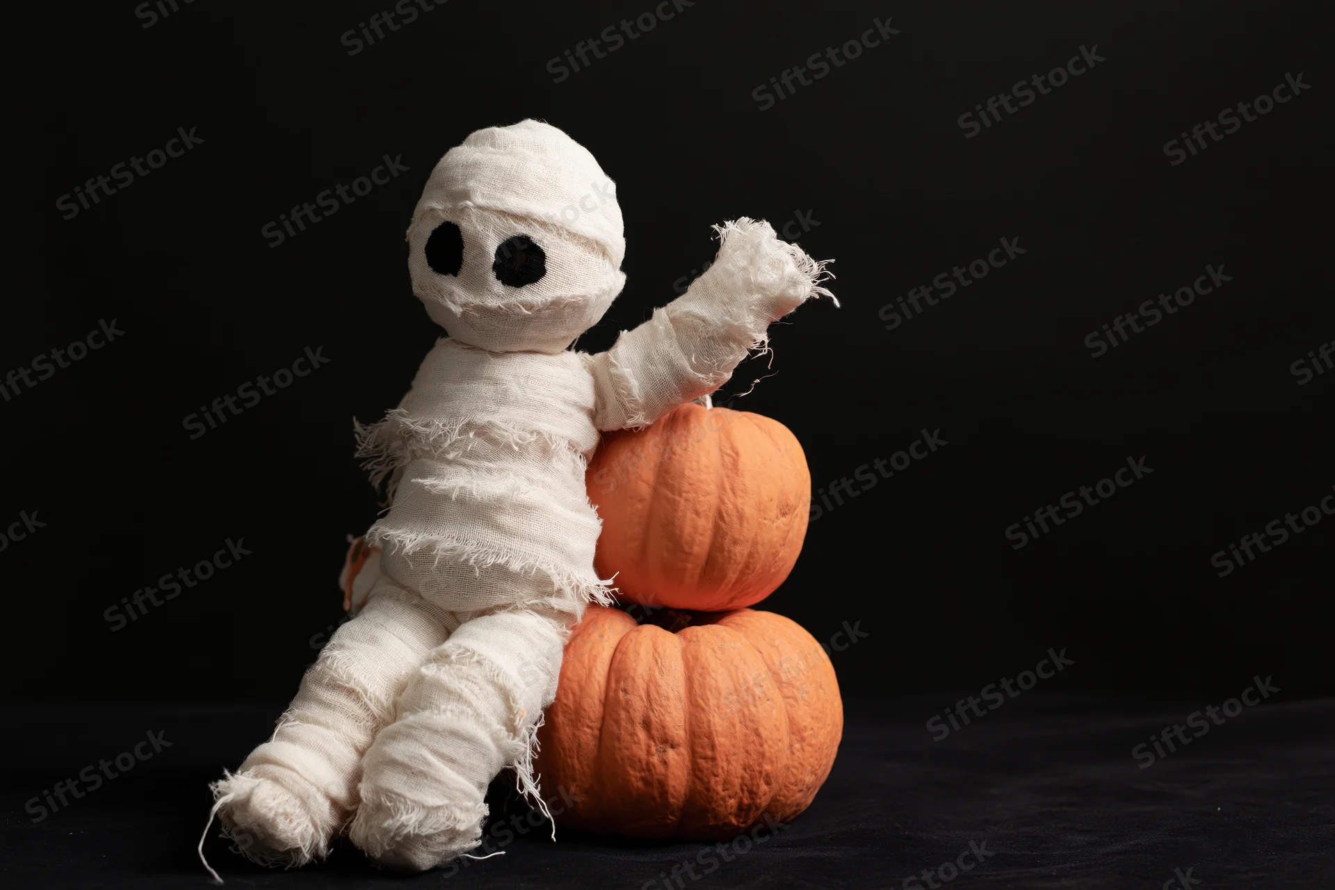 Handmade mummy figurine wrapped in white gauze bandages leaning against stacked pumpkins on black background