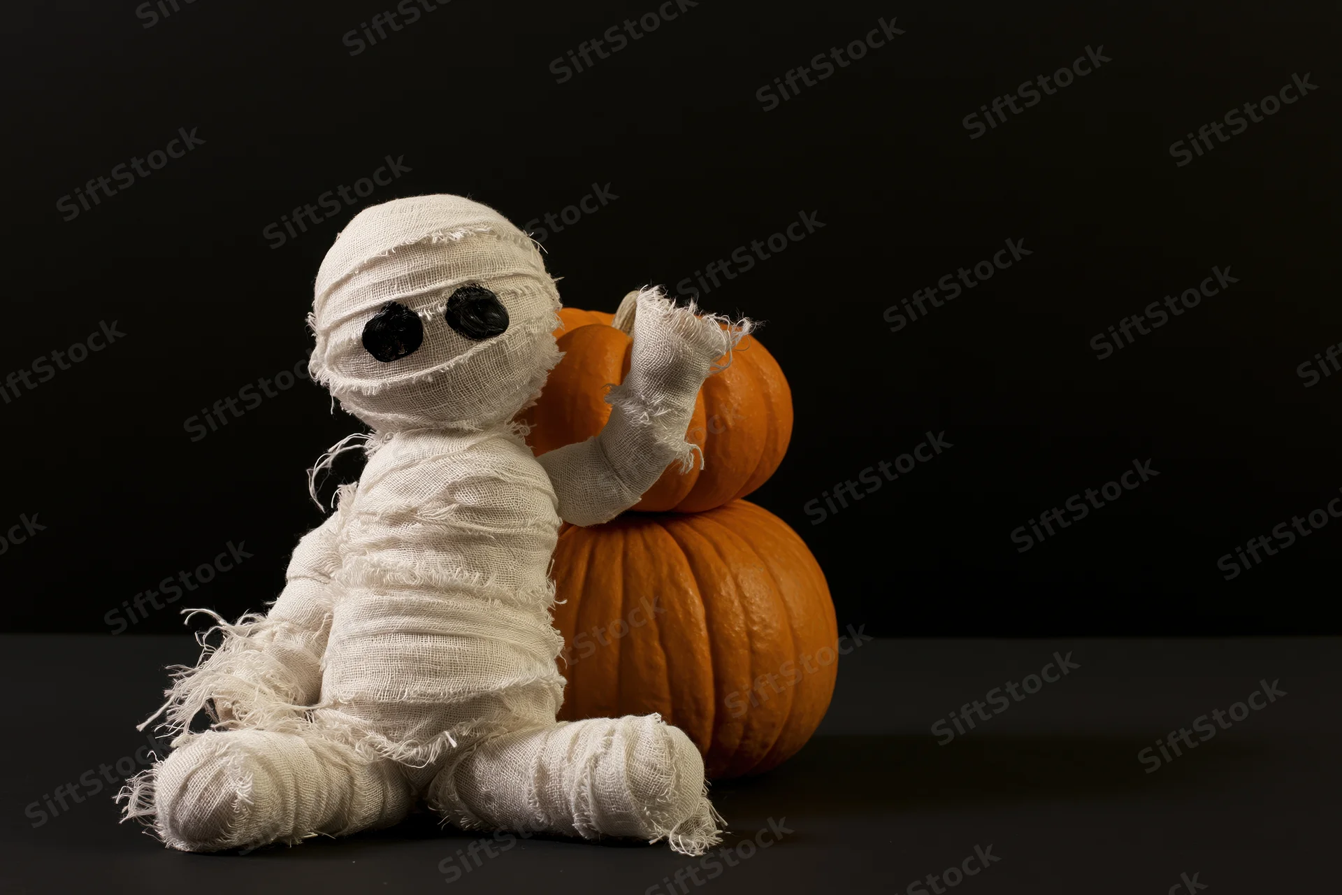 Handmade mummy figurine wrapped in white gauze bandages sitting beside stacked pumpkins on black background