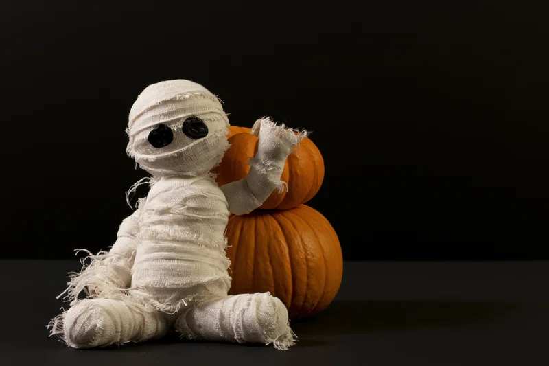 Handmade mummy figurine wrapped in white gauze bandages sitting beside stacked pumpkins on black background