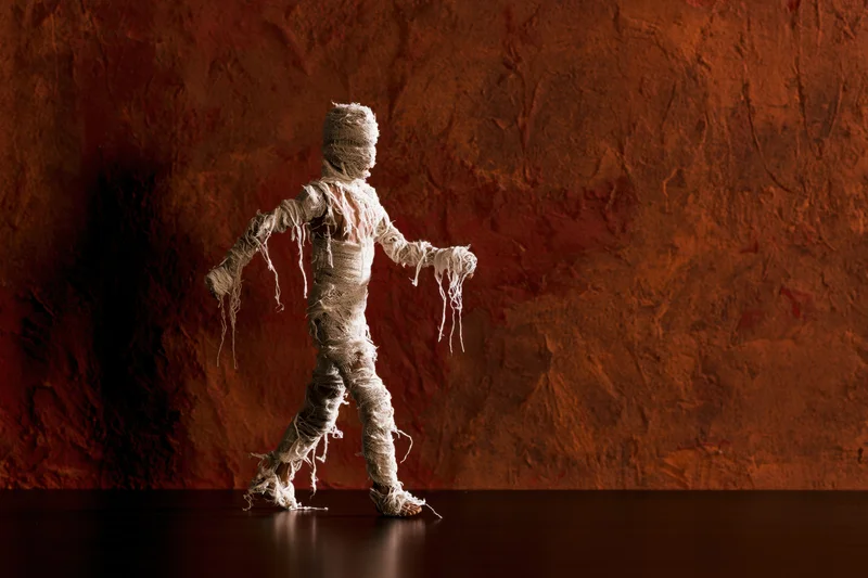 Small mummy figurine wrapped in frayed white gauze bandages walking against a textured brown wall