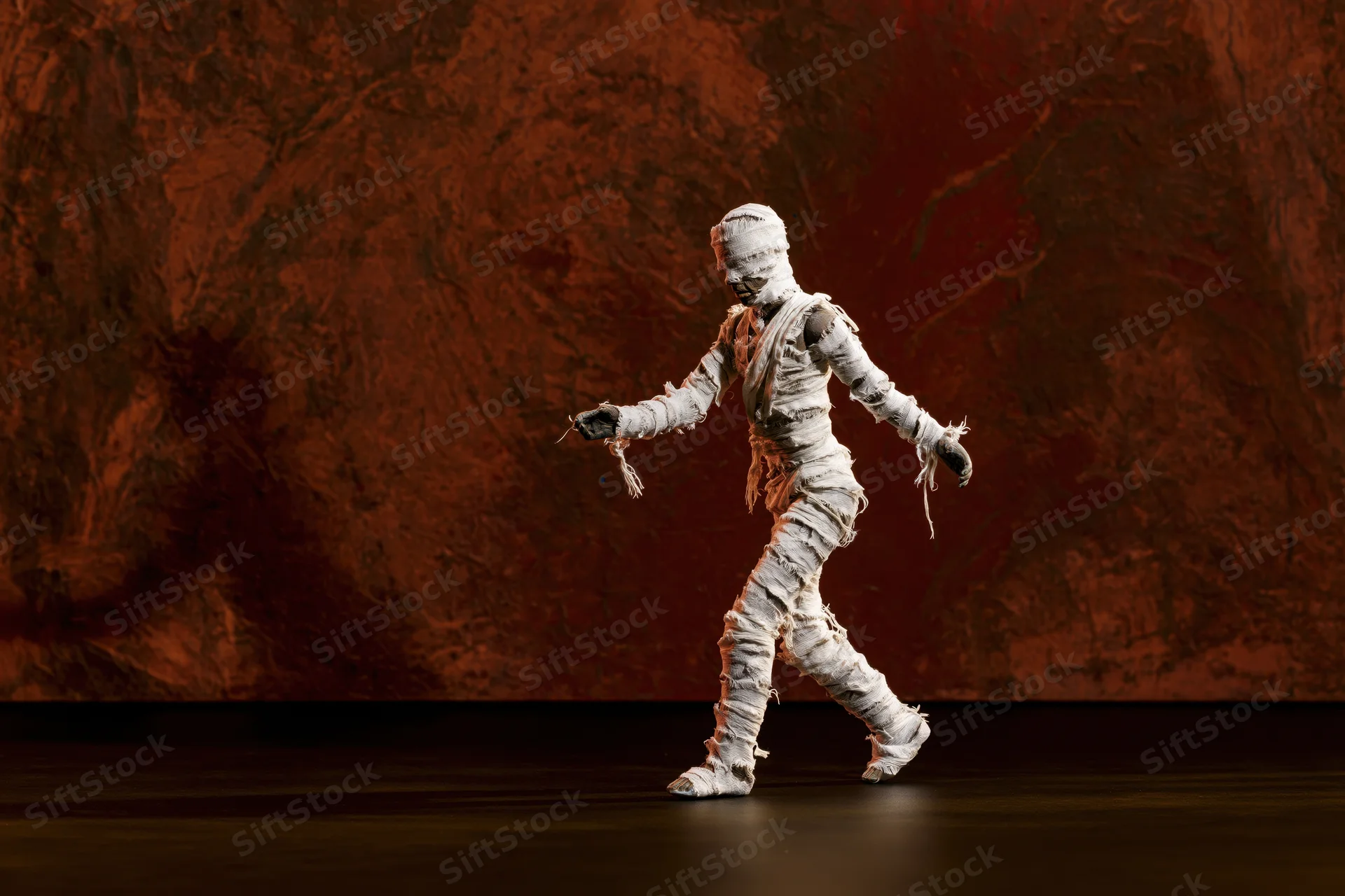 Small mummy figurine wrapped in frayed white gauze bandages walking on stage against red textured backdrop