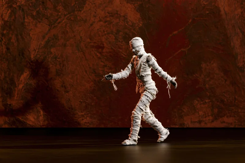 Small mummy figurine wrapped in frayed white gauze bandages walking on stage against red textured backdrop