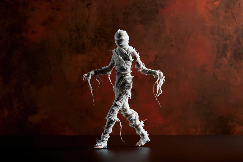 Small mummy figurine wrapped in frayed white gauze bandages walking in profile against a dark red textured wall