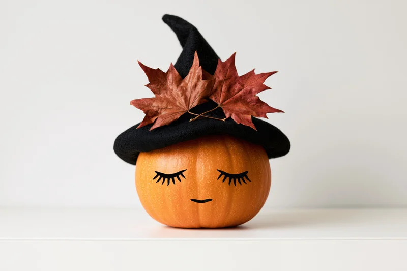 Small orange pumpkin with painted closed eyelashes wearing a black witch hat adorned with dried maple leaves on a white surface