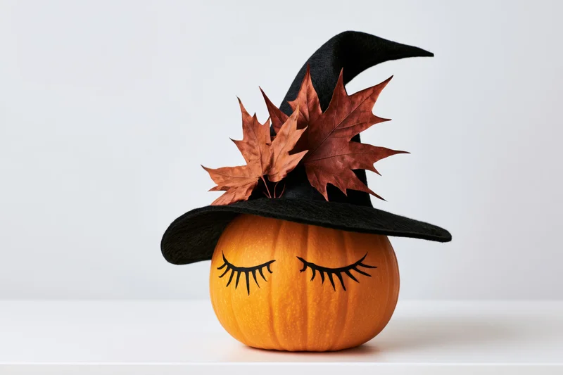 Small orange pumpkin with painted closed eyelashes wearing a black witch hat adorned with autumn maple leaves on a white surface
