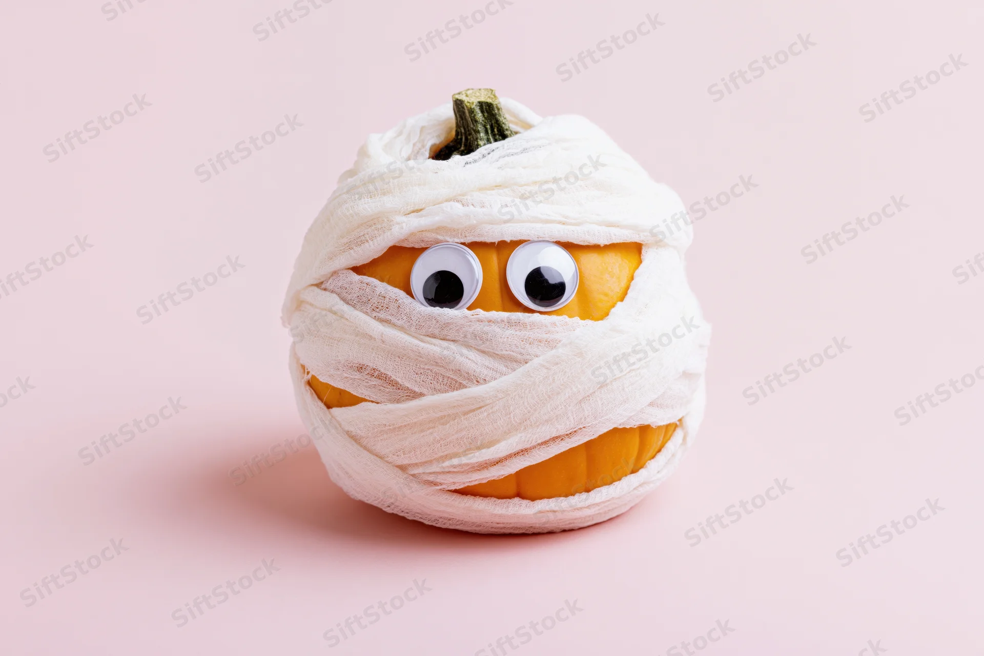 Small orange pumpkin wrapped in white gauze bandages with googly eyes on pink background