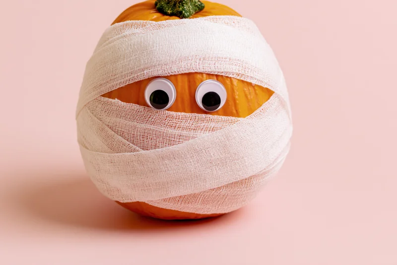 Small orange pumpkin wrapped in white gauze bandages with googly eyes on a plain pastel background