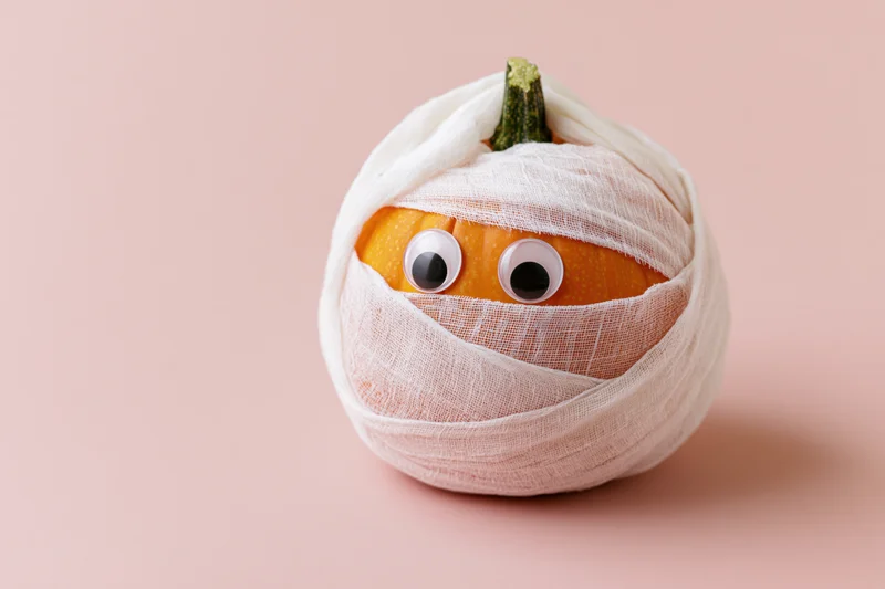 Small orange pumpkin wrapped in white gauze bandages with googly eyes on pink background