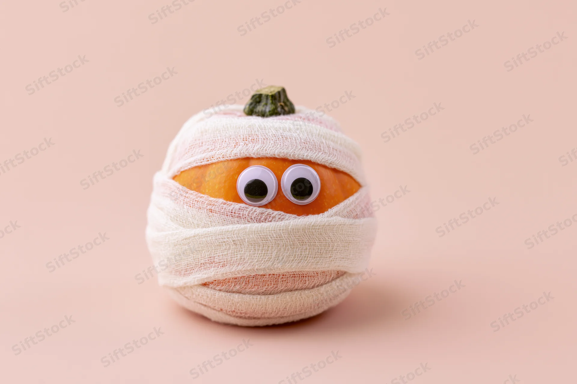 Small orange pumpkin wrapped in white gauze bandages with googly eyes on a beige background