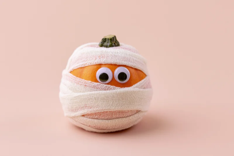 Small orange pumpkin wrapped in white gauze bandages with googly eyes on a beige background