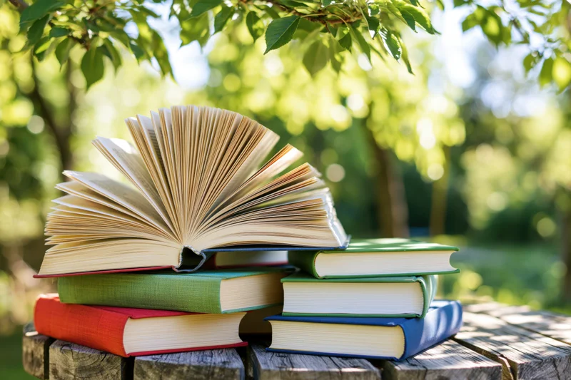 Stack of open and closed hardcover books on a weathered wooden table outdoors under tree leaves