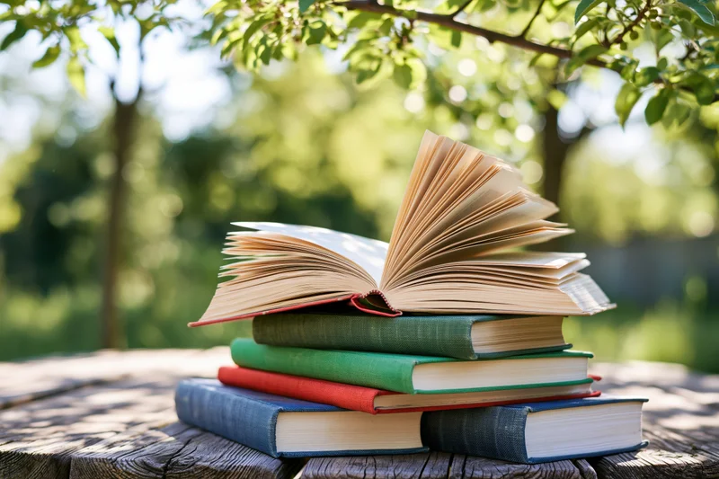 Stack of open and closed hardcover books on a weathered wooden table outdoors under tree leaves