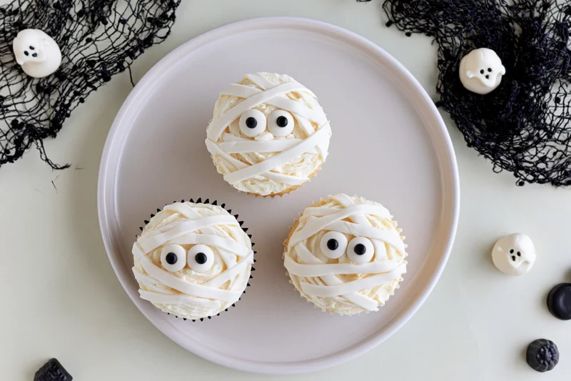Three halloween mummy cupcakes with candy eyes and white frosting on a plate surrounded by black netting and ghost decorations