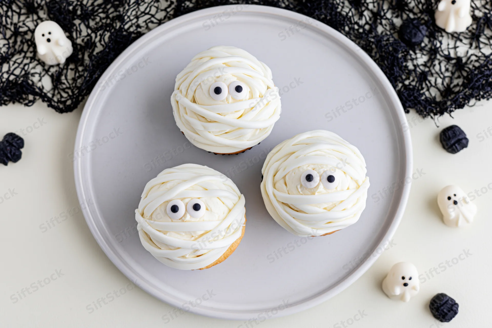 Three halloween mummy cupcakes with candy eyes on a white plate surrounded by ghost decorations and black lace