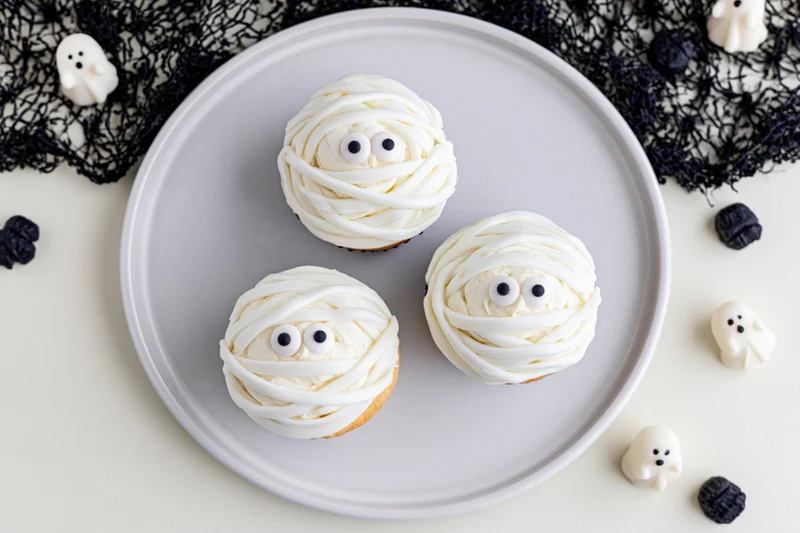 Three halloween mummy cupcakes with candy eyes on a white plate surrounded by ghost decorations and black lace