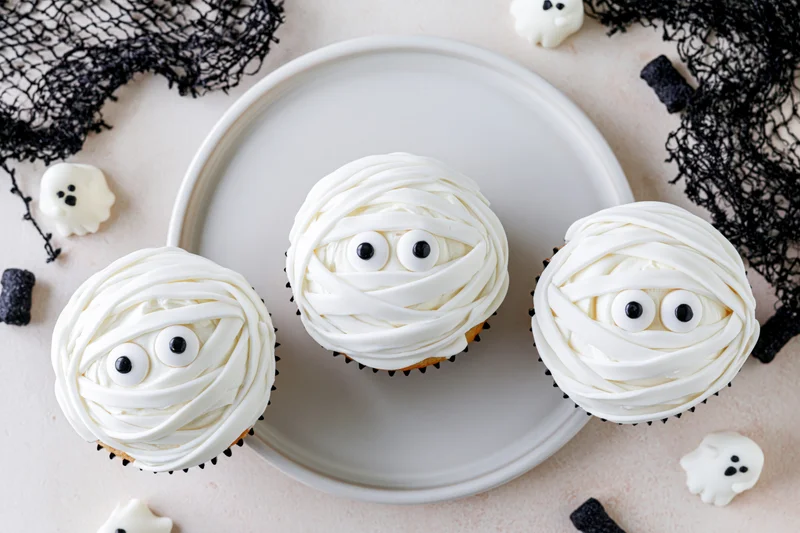 Three halloween mummy cupcakes with white frosting and candy eyes on a ceramic plate surrounded by ghost decorations and black netting