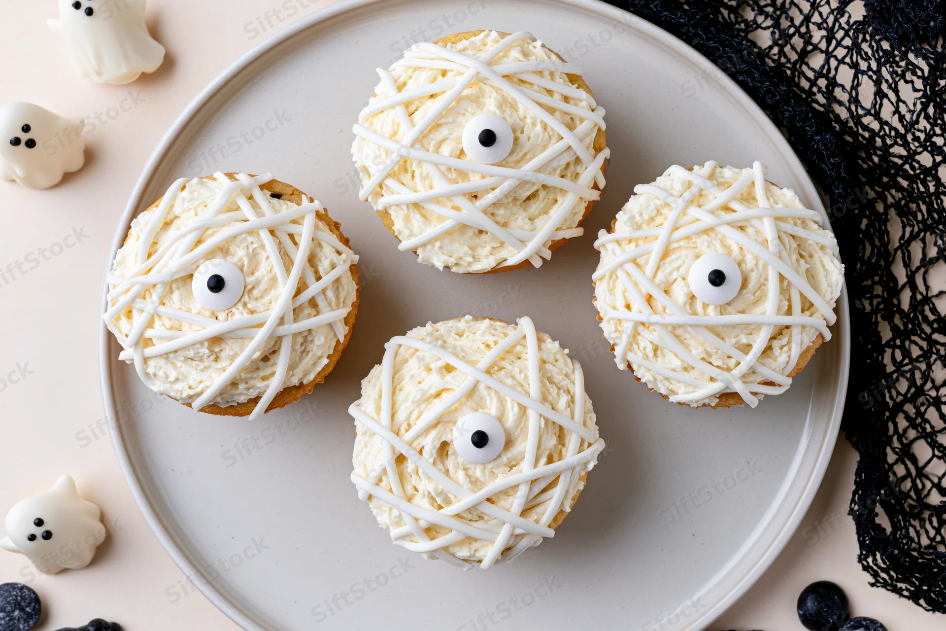 Four mummy-themed cupcakes with white frosting and candy eyes on a ceramic plate surrounded by Halloween decorations