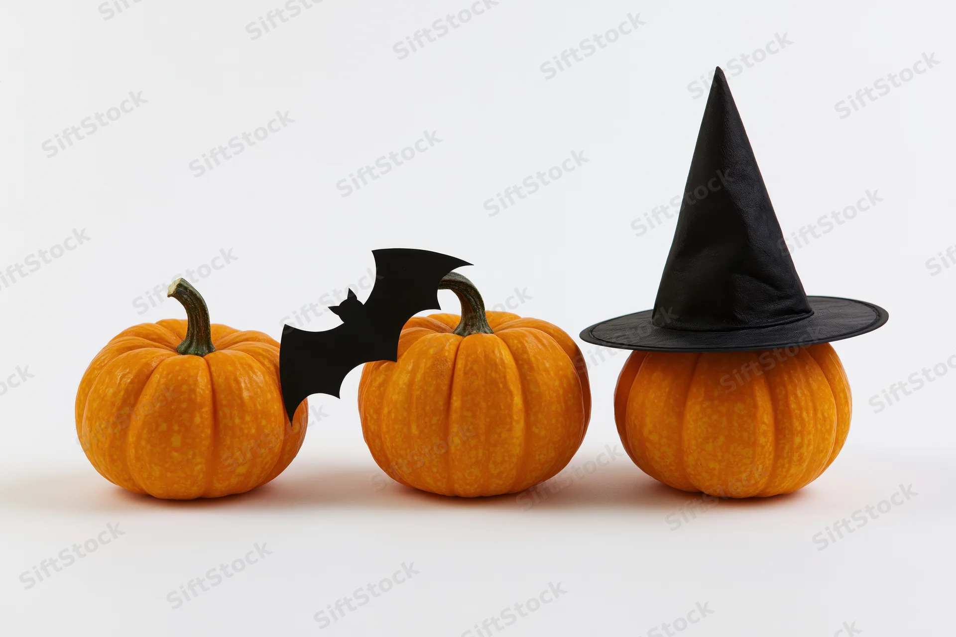 Three small orange pumpkins in a row, one wearing a witch hat and another with a bat cutout, on a white background