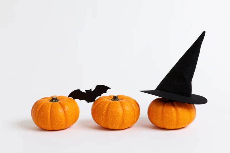 Three small orange pumpkins in a row with a witch hat and bat cutout on white background