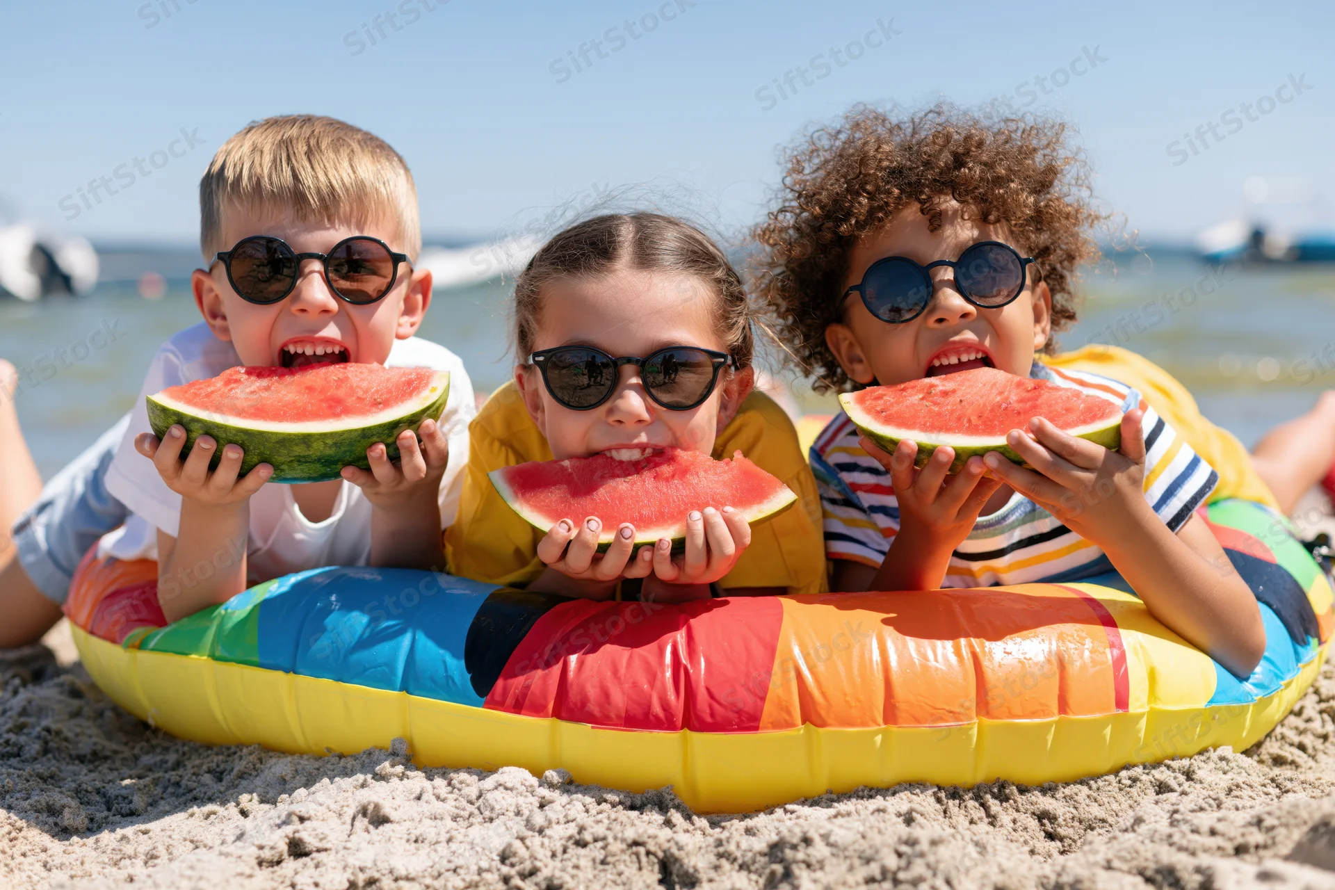 Three children lying on a colorful inflatable pool float on the beach eating watermelon