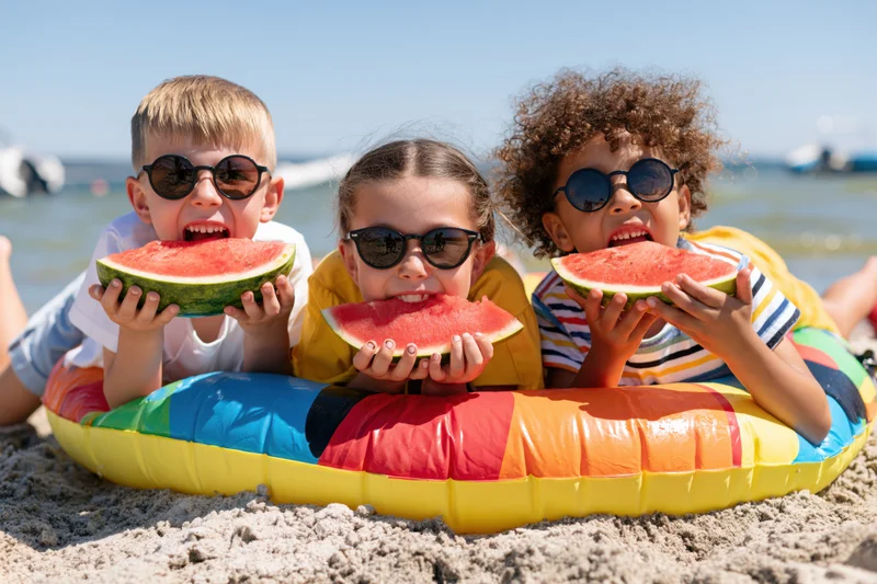 Three children lying on a colorful inflatable pool float on the beach eating watermelon