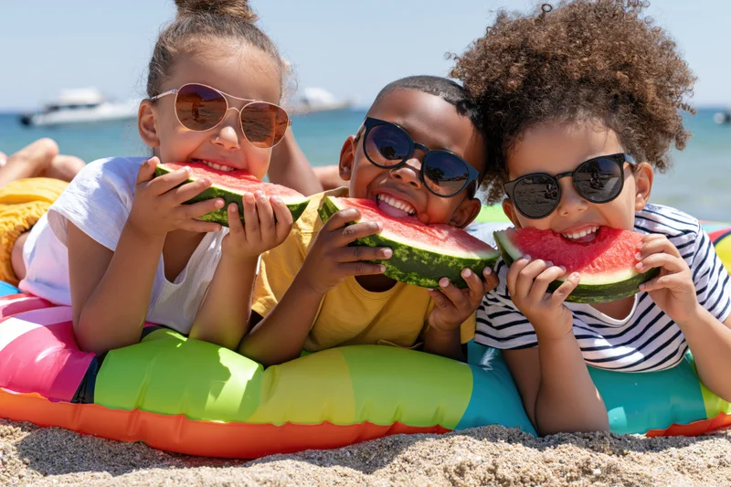 Three young children lying on a colorful inflatable float on the beach eating watermelon while wearing sunglasses
