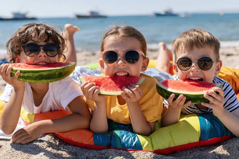 Three children lying on a colorful inflatable float on the beach eating watermelon while wearing sunglasses