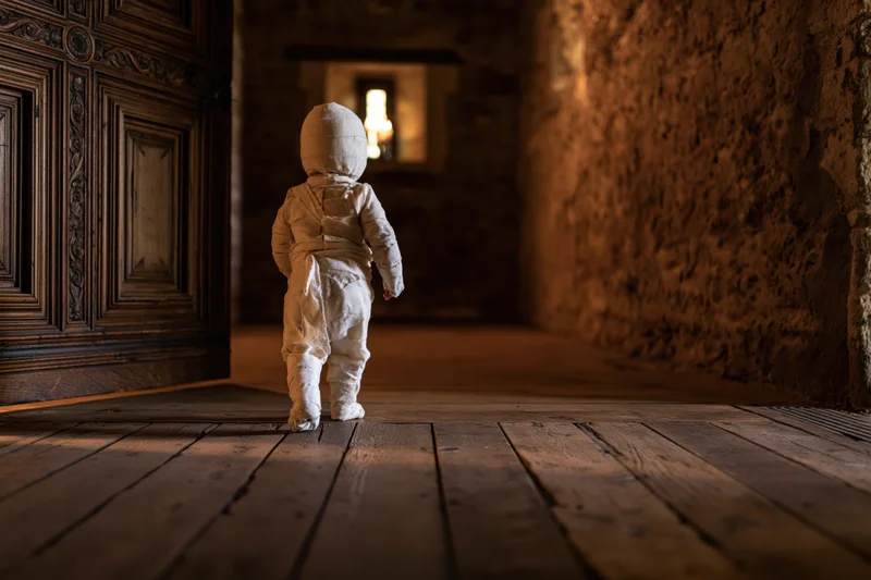 Toddler in white bandage mummy costume walking down dim stone hallway toward light