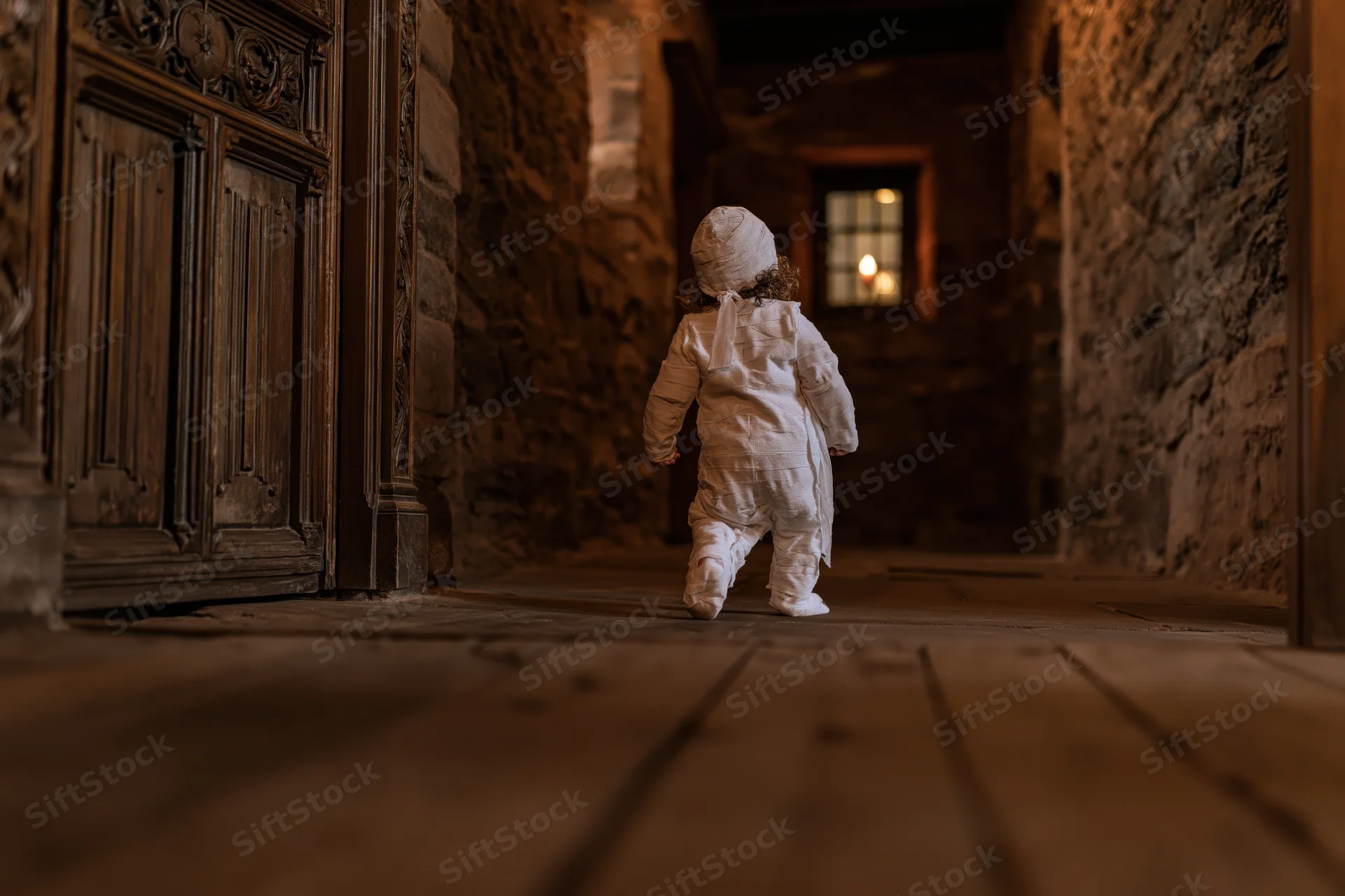 Toddler in white mummy costume walking down dim stone hallway toward candlelit window