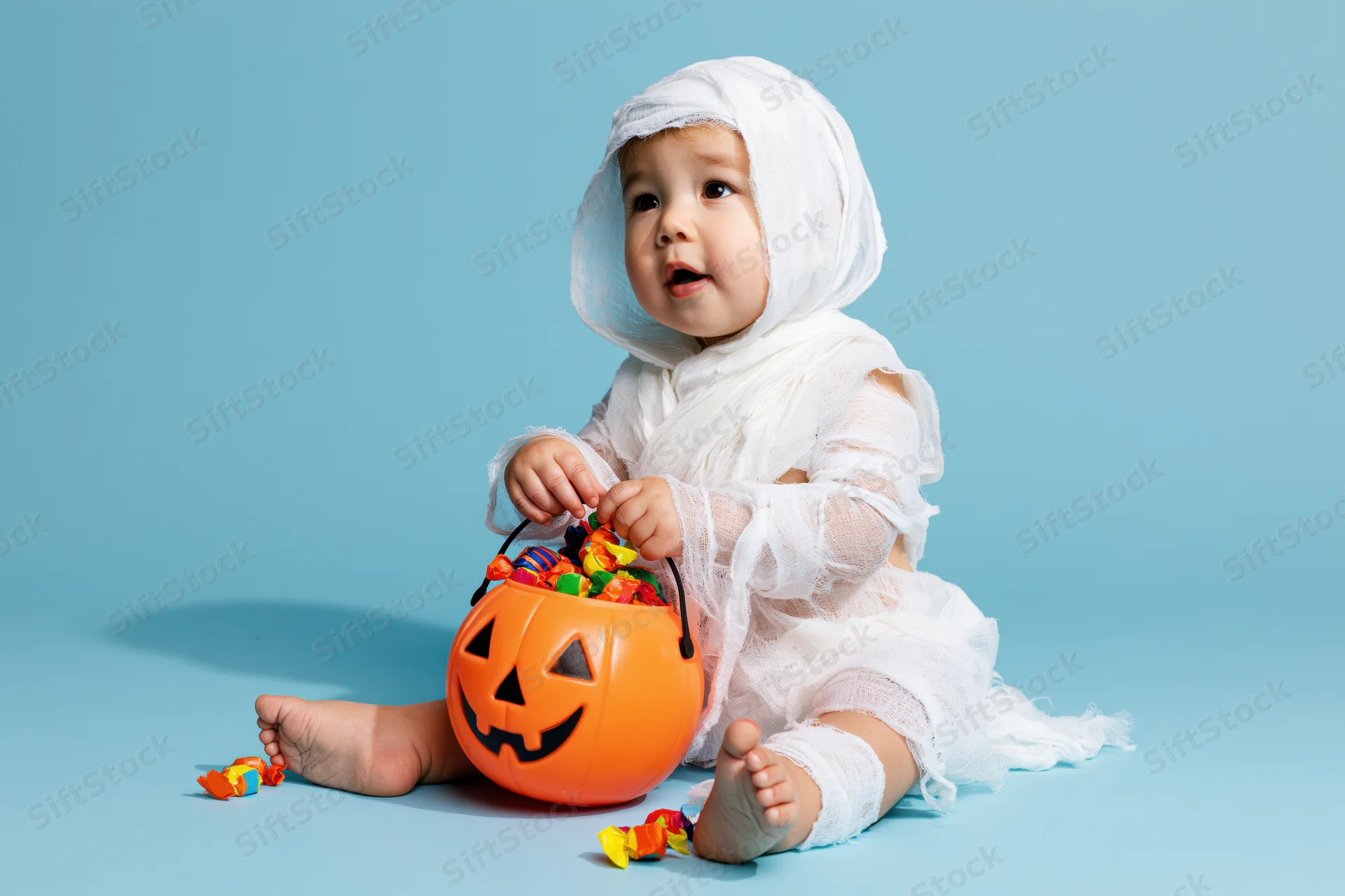 Toddler in white gauze mummy costume sitting with orange jack-o-lantern candy bucket on blue background