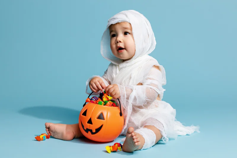 Toddler in white gauze mummy costume sitting with orange jack-o-lantern candy bucket on blue background