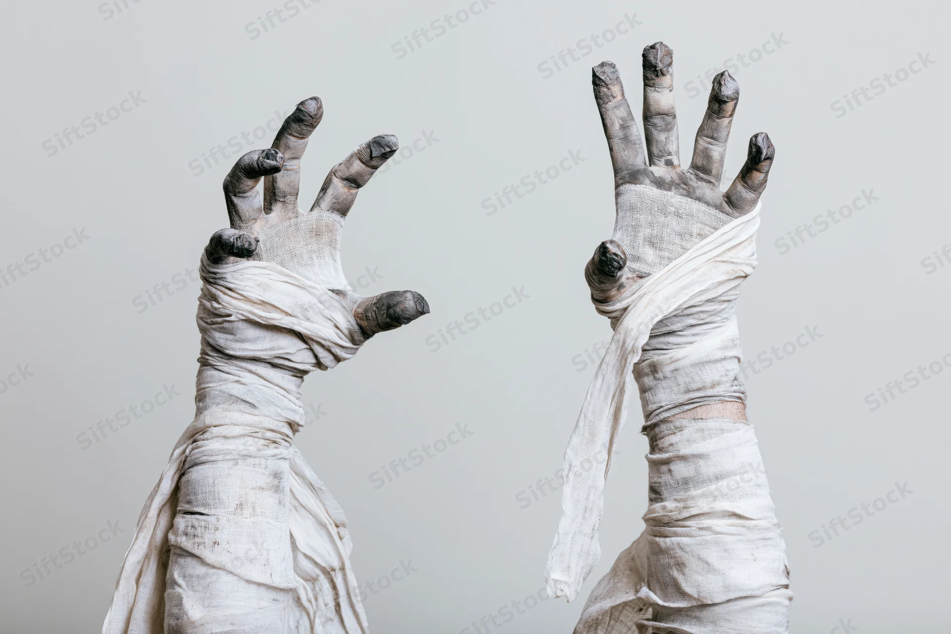 Two mummy hands wrapped in linen bandages raised with fingers spread in a clawing gesture against a plain background