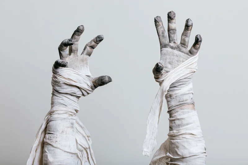 Two mummy hands wrapped in linen bandages raised with fingers spread in a clawing gesture against a plain background