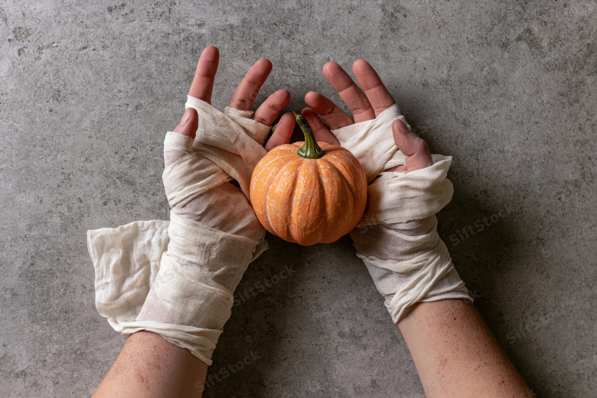 Hands wrapped in cream gauze bandages holding a small orange pumpkin on a concrete surface