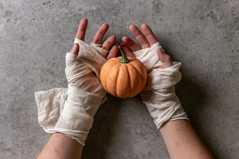 Hands wrapped in cream gauze bandages holding a small orange pumpkin on a concrete surface