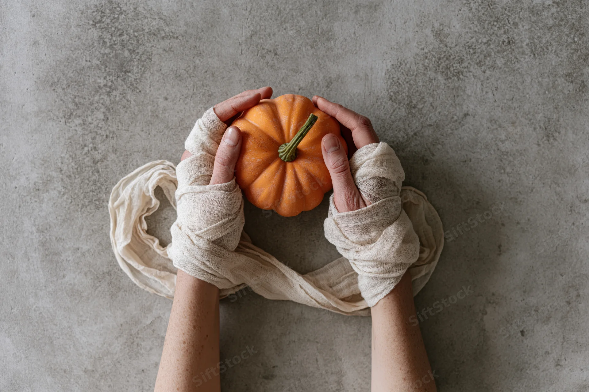 Hands wrapped in cream gauze bandages cradling a small orange pumpkin on a gray concrete surface