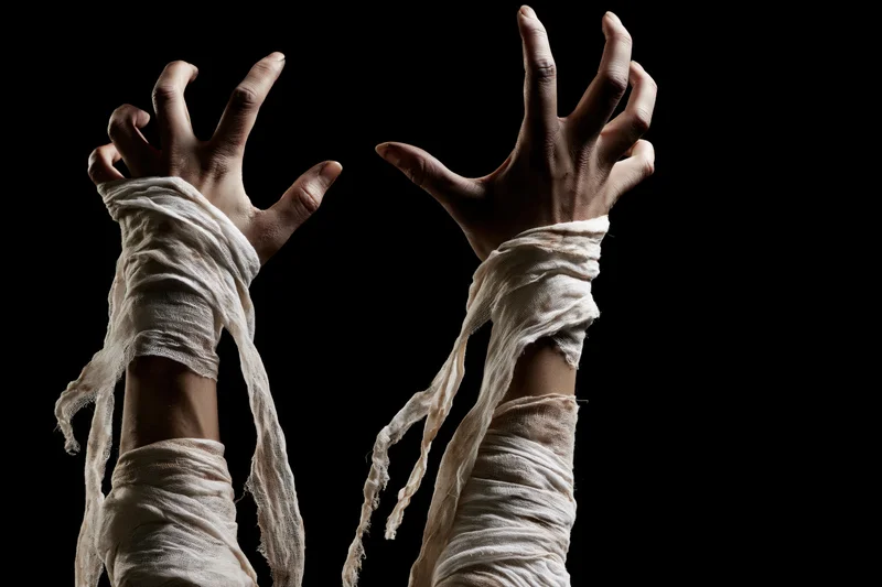 Mummy hands wrapped in tattered bandages reaching upward against black background
