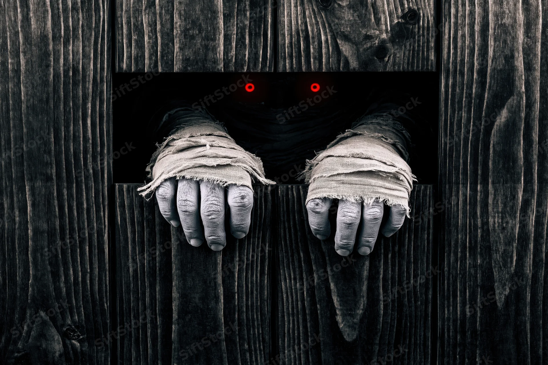 Mummy hands wrapped in bandages gripping a wooden door with glowing red eyes visible through a gap