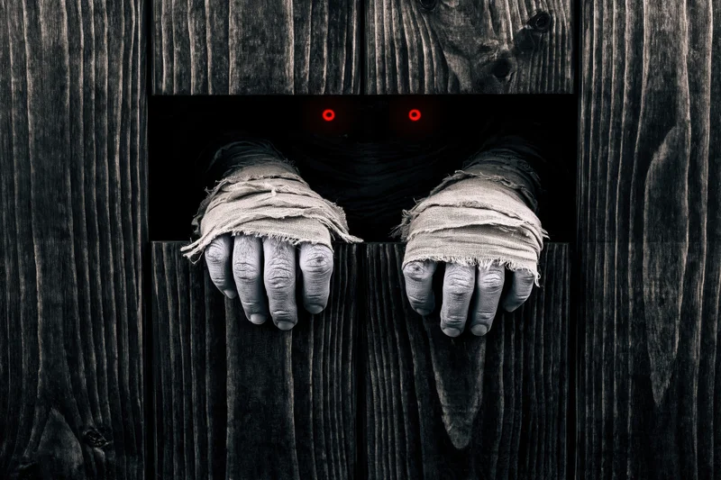 Mummy hands wrapped in bandages gripping a wooden door with glowing red eyes visible through a gap