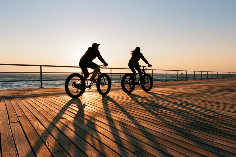 Two people riding bicycles side by side on a wooden boardwalk at sunset by the ocean
