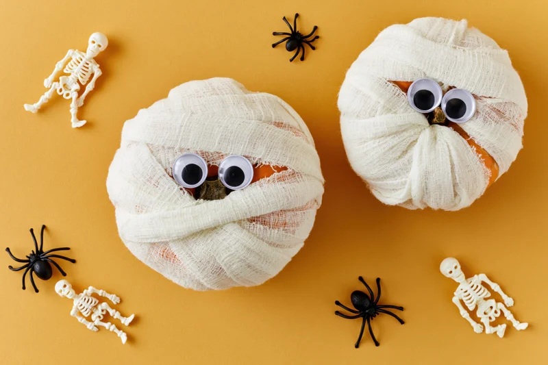 Two pumpkins wrapped in white gauze with googly eyes surrounded by toy skeletons and spiders on orange background