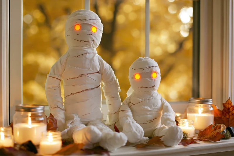 Two white fabric mummy stuffed toys with glowing orange eyes on a windowsill surrounded by candles and autumn leaves