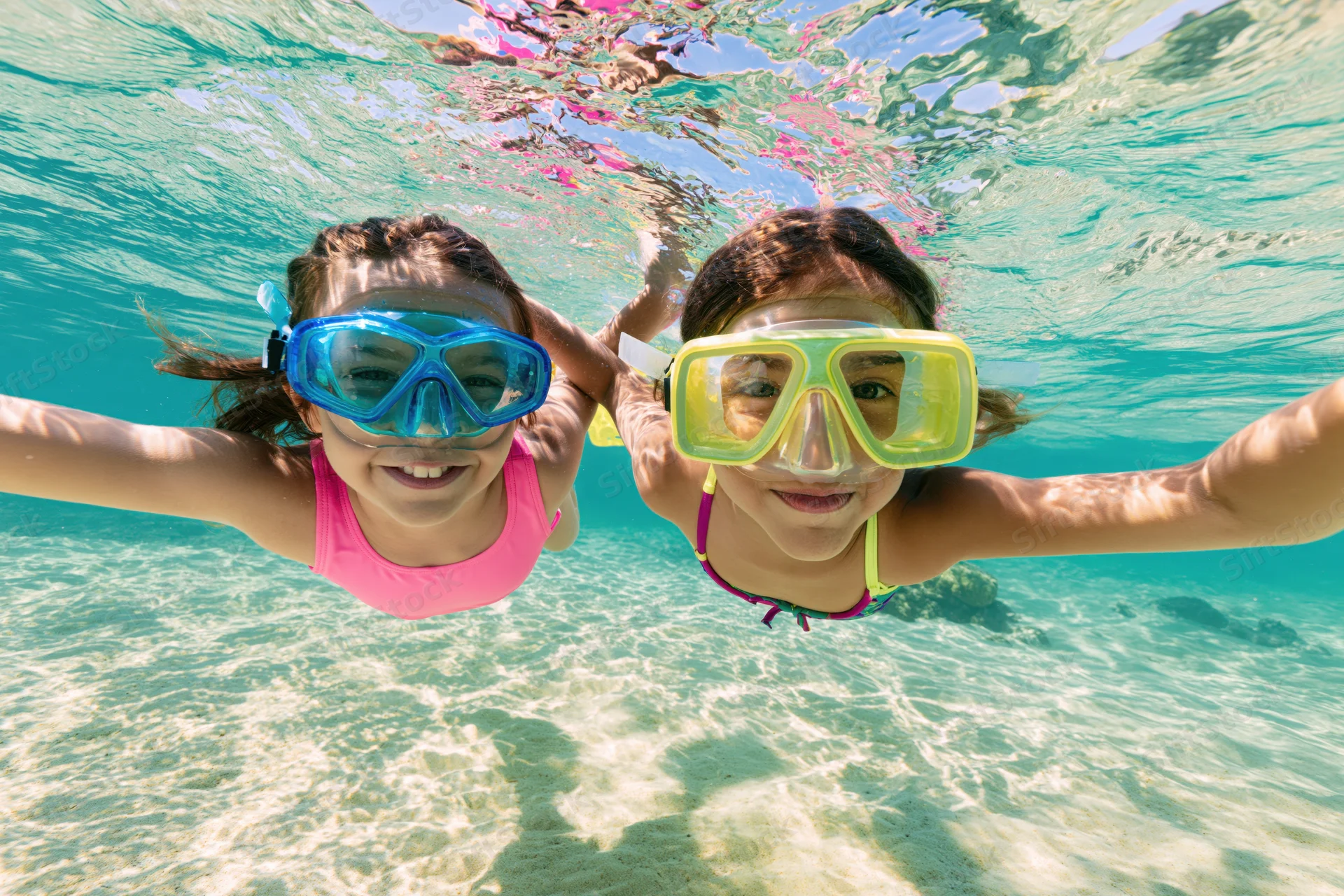 Two young girls swimming underwater in shallow tropical sea wearing snorkel masks and smiling