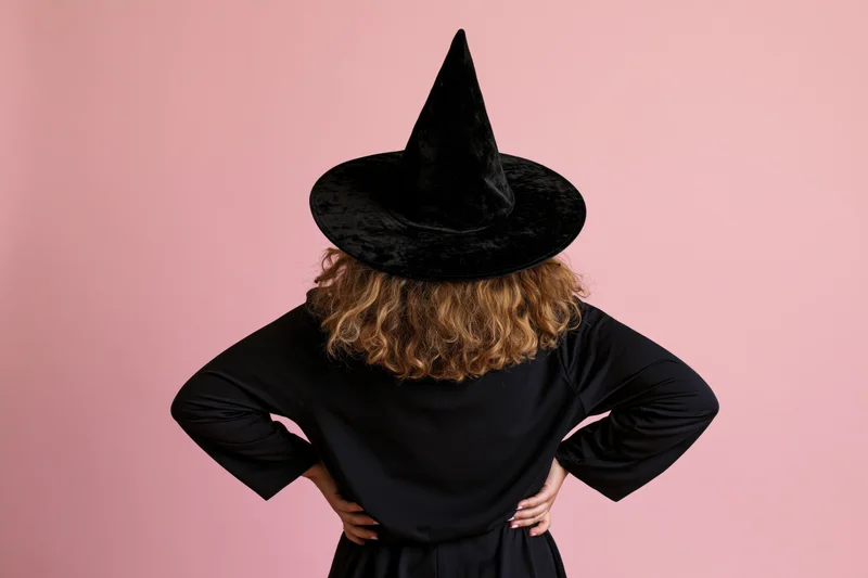 Woman in witch costume with hands on hips seen from back against pink background