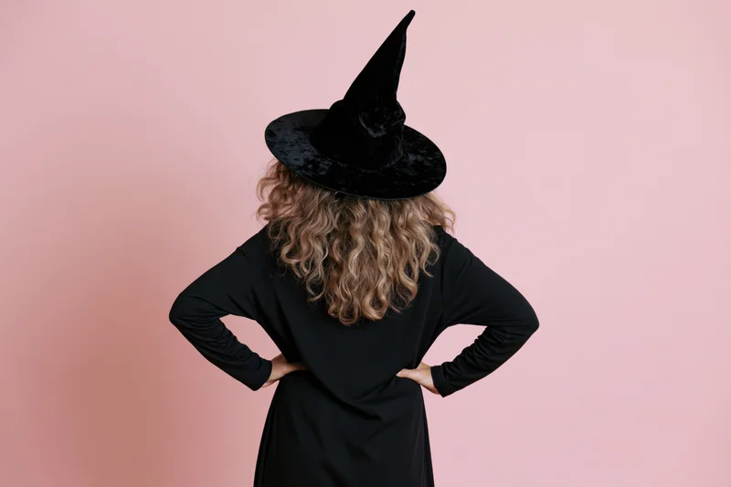 Woman in witch costume with hands on hips seen from back against pink background