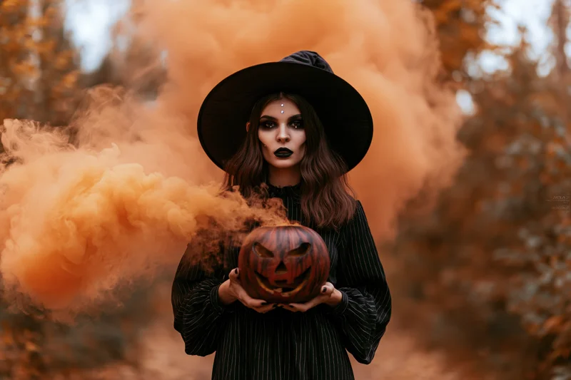 Woman in witch costume holding carved pumpkin amidst orange smoke in forest