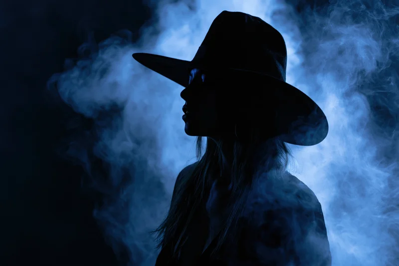 Woman in wide-brimmed witch hat in silhouette against blue smoke at night