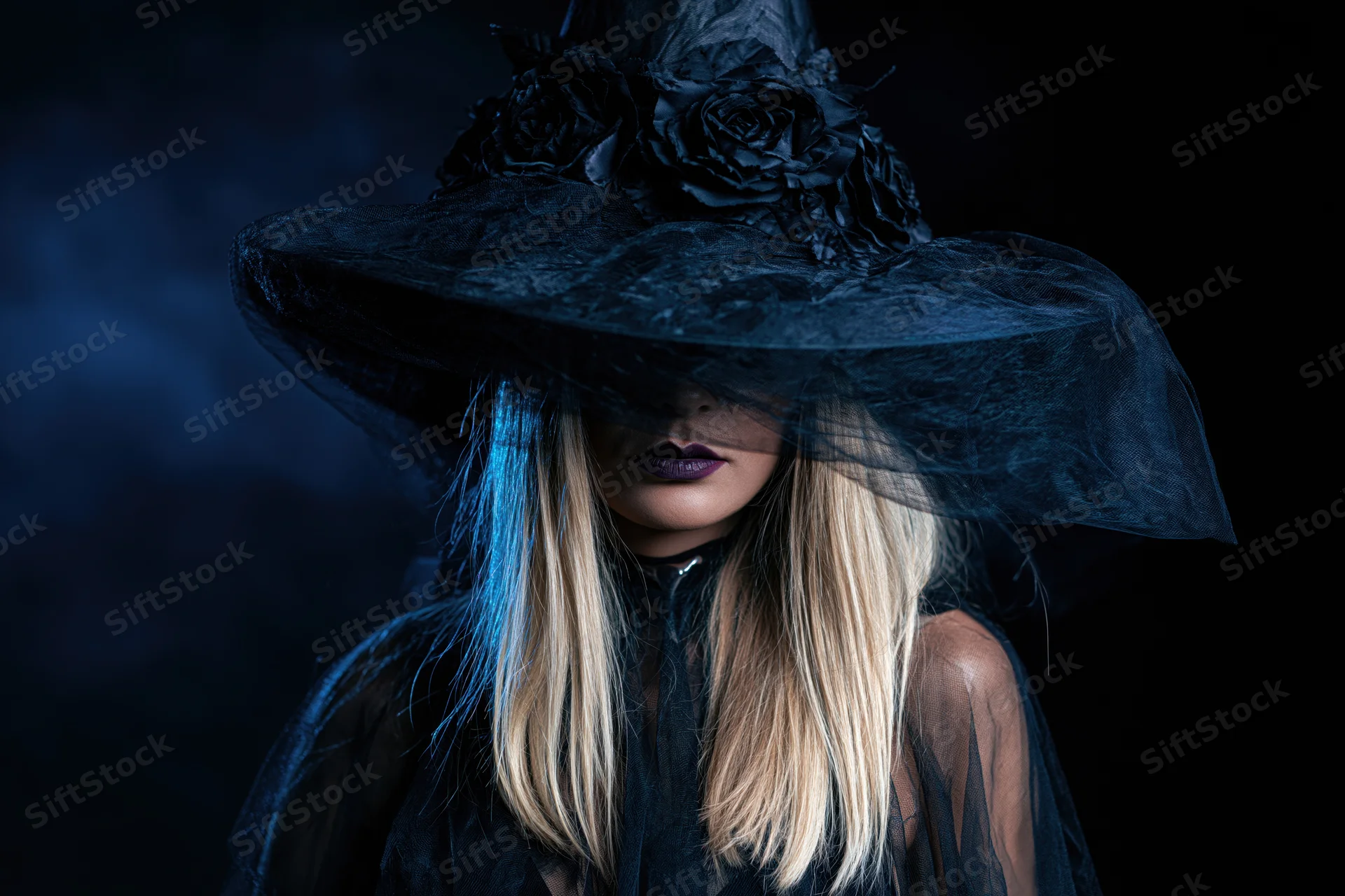 Woman in black witch hat with dark roses and veil, blonde hair, dark lipstick, close-up portrait against dark background