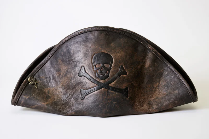 Worn leather pirate tricorn hat with skull and crossbones emblem displayed flat on white background
